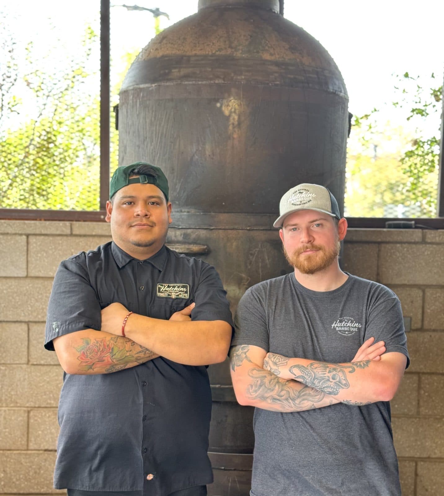 two men crossing arms in front of bbq smoker and brick wall